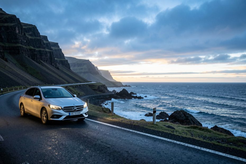 Une voiture de location argentée roule sur une route côtière islandaise au lever du jour, entre des montagnes sombres sous un ciel changeant et une lumière froide.